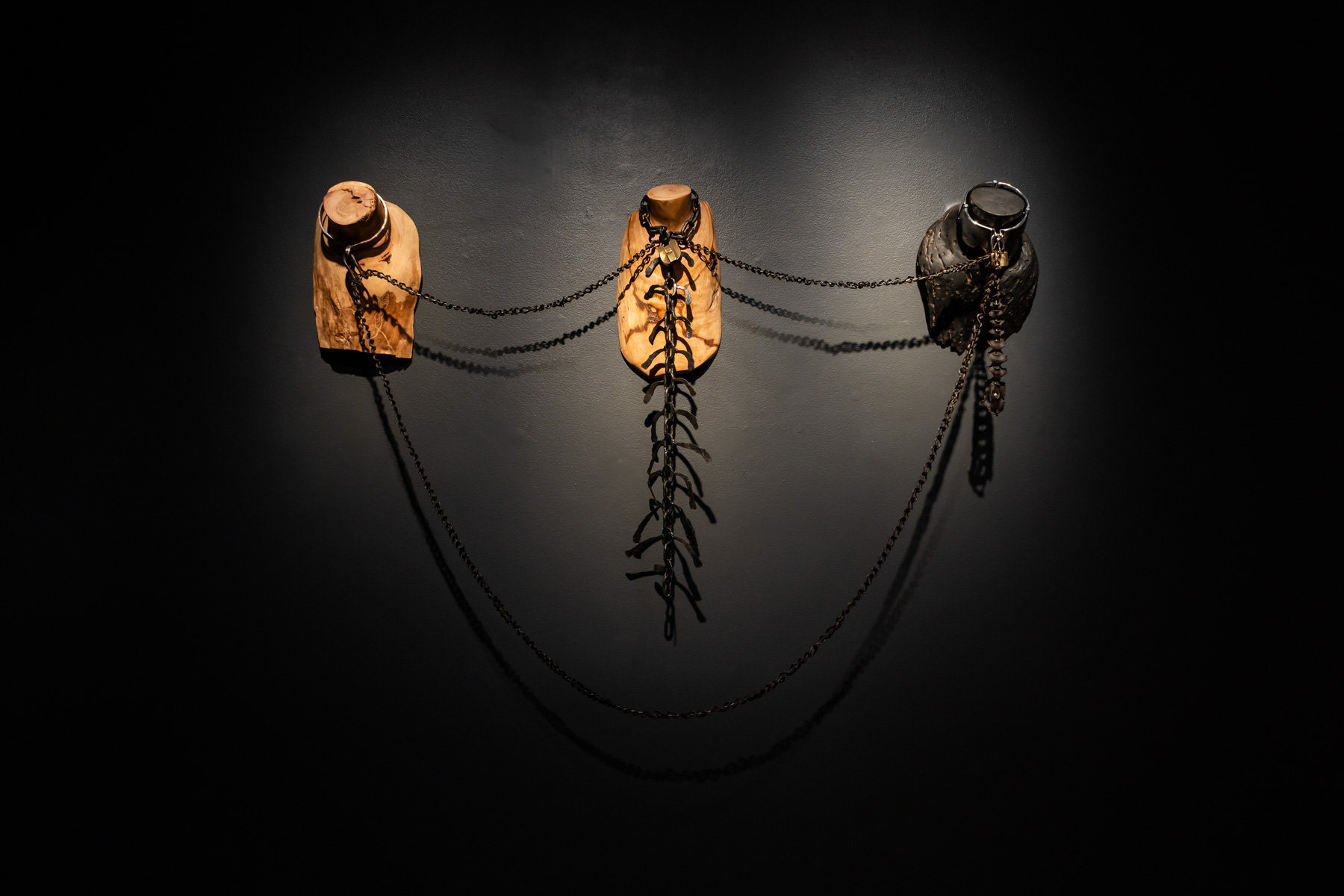 Three wooden busts, displaying neck and torso only, connected by a chain and mounted directly onto a black wall