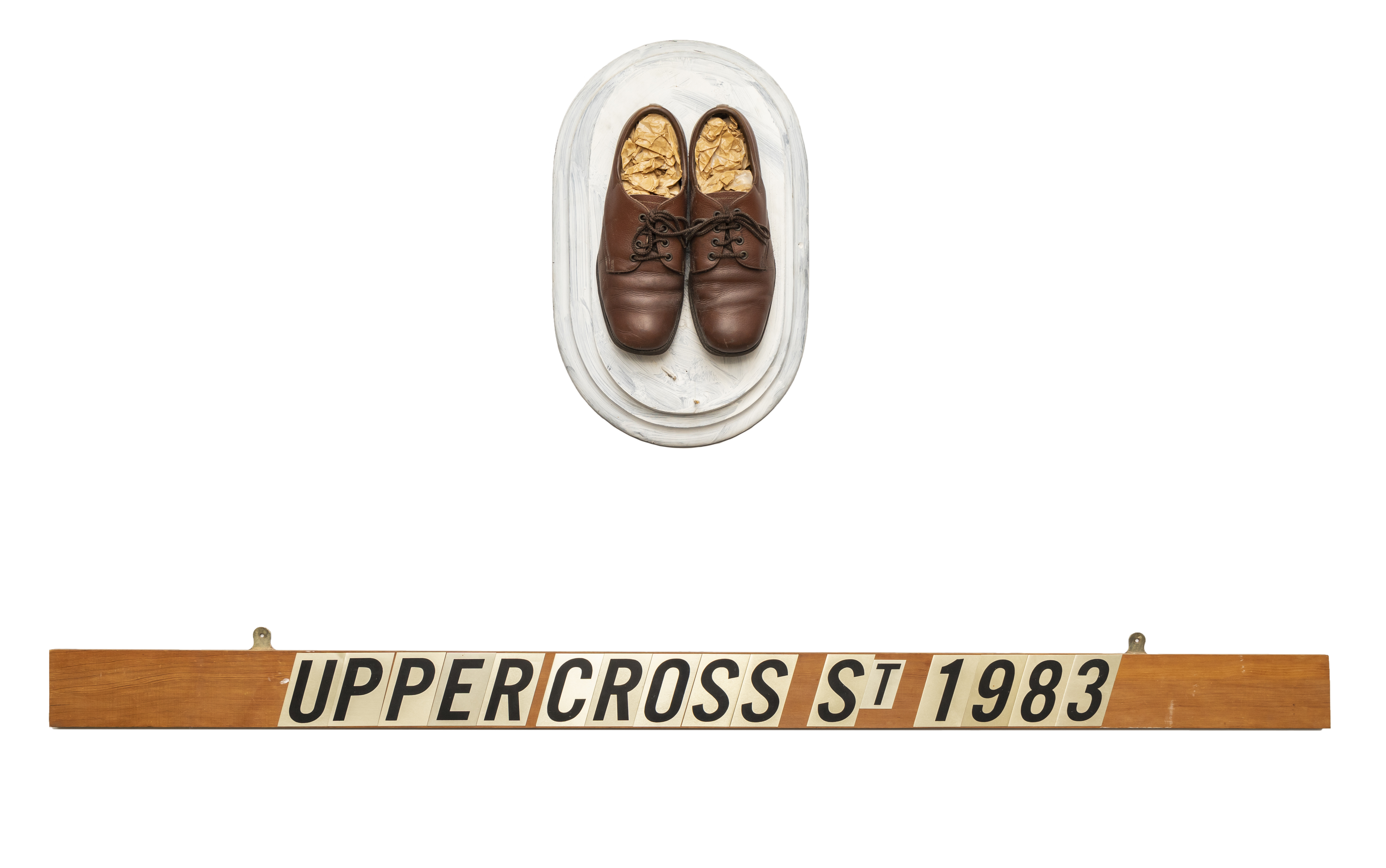 A pair of brown leather dress shoes on white wooden mount. Below the shoes is a wooden plank with lettering that reads 'Upper Cross St 1983'.