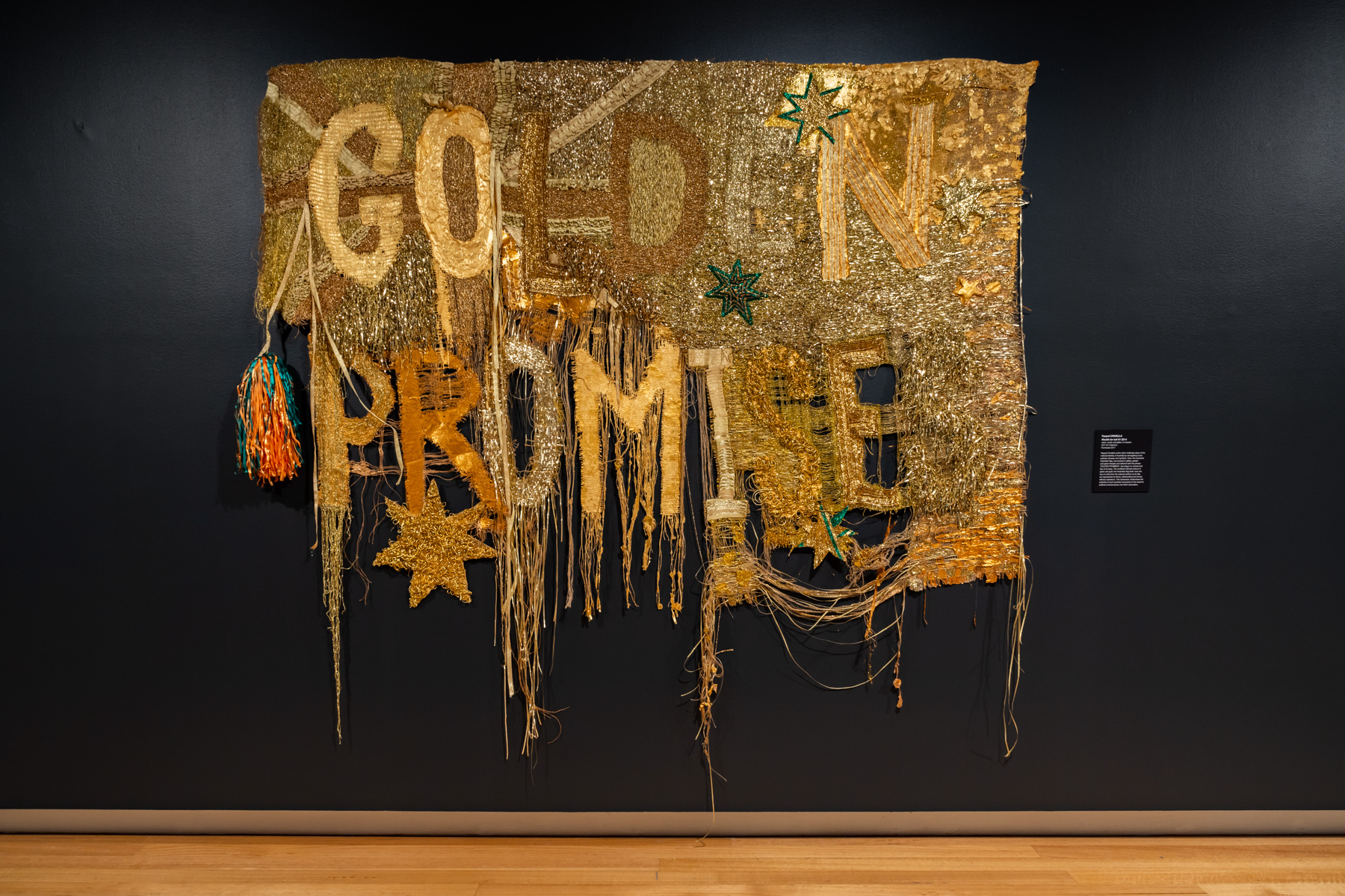 A golden woven Australian flag featuring the words ‘Golden Promises.’ In the bottom left-hand corner, the flag has frayed away leaving only the text and star motifs.