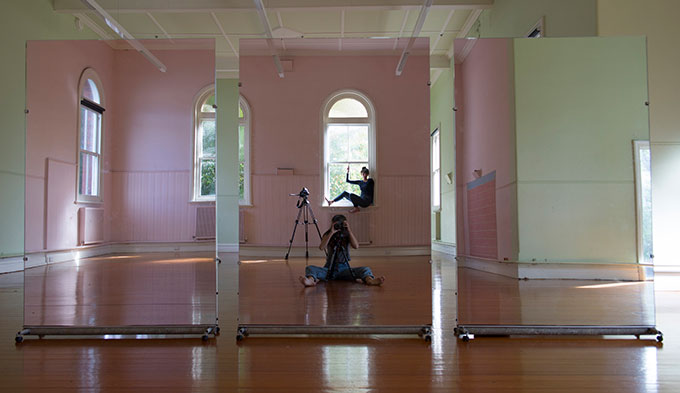 Image of two women in a room looking at their mirrored reflection. One woman in window and one woman sitting on ground with a camera 