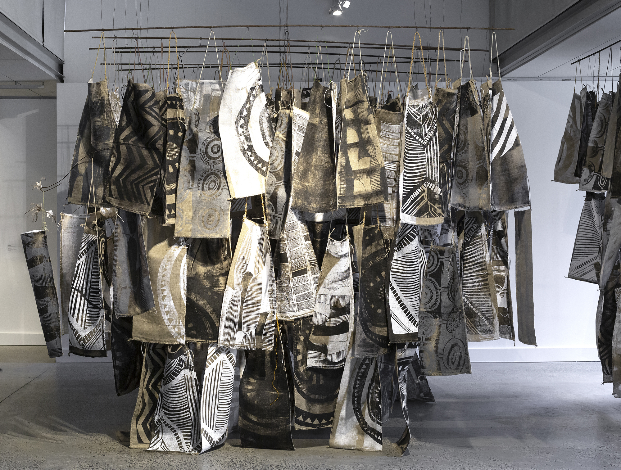 Dozens of pieces of hessian-coloured fabric printed with black-and-white geometric designs and suspended in a tight cluster in an art gallery.