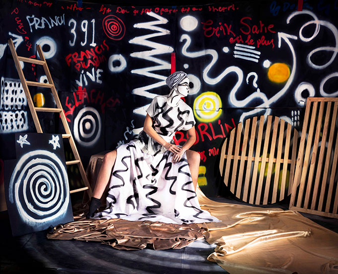 Image of painted white painted lady with white dress sitting in front of a black wall with graffiti with wooden ladder and other objects on a black background